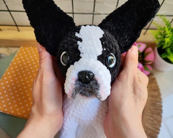 May include: A handmade plush Boston Terrier dog with large black ears, a black nose, and a white and black face. The dog is held by two hands. The plush dog is a soft, cuddly toy.