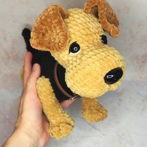 May include: A crocheted plush toy dog with brown ears, black eyes, and a black nose. The dog is wearing a black collar with a silver buckle. The dog is sitting on a person's hand.