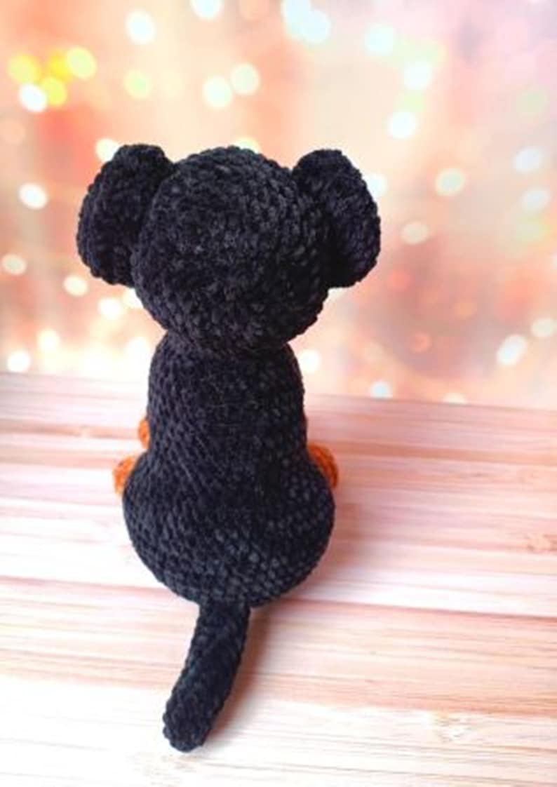 May include: Black crocheted plush toy dog with orange feet and a long tail. The dog is sitting on a wooden surface.
