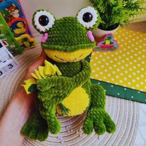 May include: A handmade green and yellow crochet frog with large white-rimmed eyes and a yellow belly. The frog is holding a bouquet of yellow flowers. The frog is a plush toy.