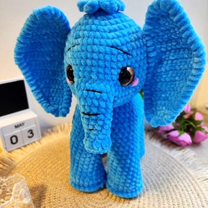 Może przedstawiać: Ręcznie robiony, pluszowy niebieski słoń z dużymi uszami, trąbą i małymi kłami. Słoń ma duże, ciemne oczy i różowe policzki. Słoń stoi na tkanej macie. Kalendarz pokazuje datę 3 maja.