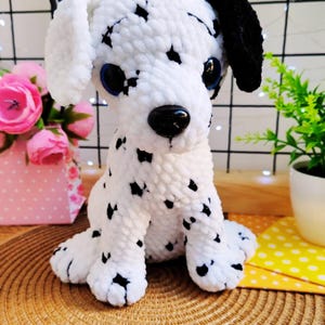 Puede incluir: Un peluche de perro dálmata hecho a mano, blanco con manchas negras, ojos azules y nariz negra. El peluche está sobre una estera tejida. Se ven flores rosas y una pequeña planta en el fondo.