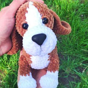 Puede incluir: Un juguete de peluche hecho a mano con hilo marrón y blanco que representa un perro. El perro tiene la cara, el pecho y las patas blancas, con las orejas y el cuerpo marrones. Tiene ojos negros y una nariz negra. El juguete se sostiene en una mano.