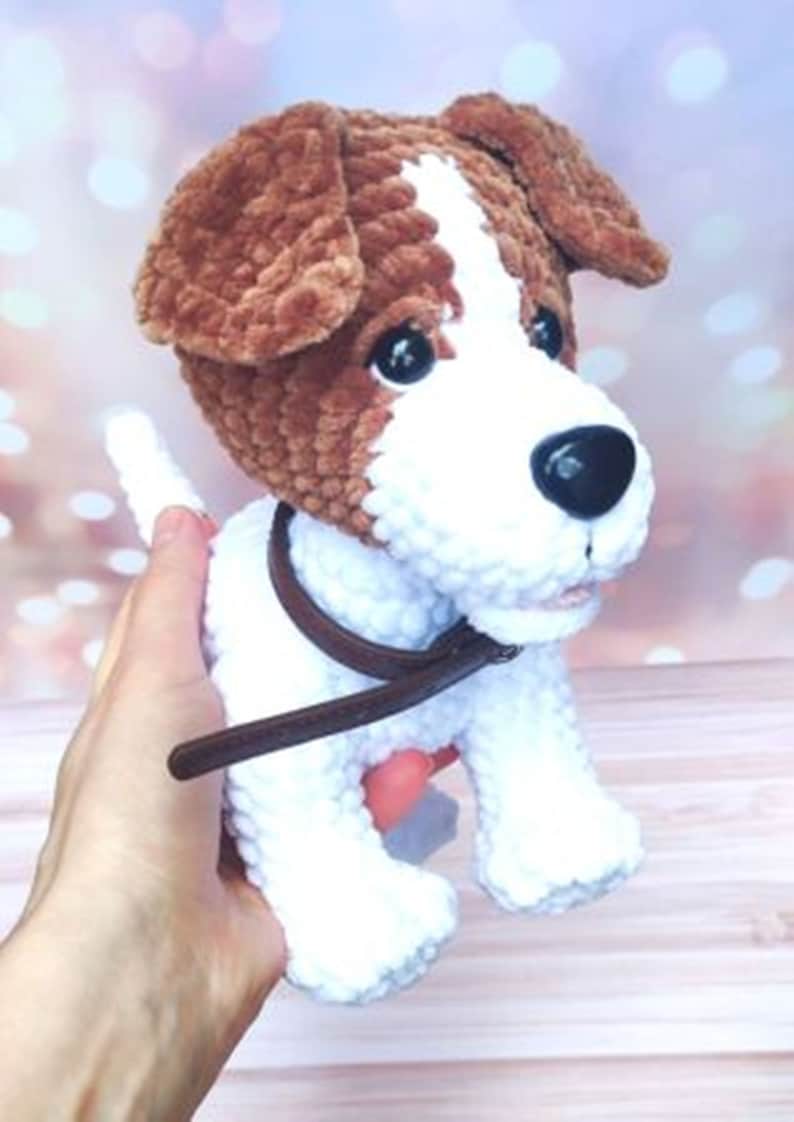 Op de afbeelding: Een wit en bruin gehaakt hondenspeelgoed met een bruin leren halsband. De hond heeft zwarte ogen en een zwarte neus.
