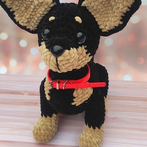 May include: A crocheted plush toy dog with black and tan fur, wearing a red collar with a silver buckle. The dog is sitting on a light brown wooden surface.