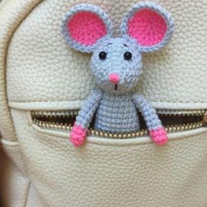 Peut inclure: Une souris crochetée, corps gris, oreilles, nez et pieds roses, sort d'une poche zippée crème. La souris a des yeux noirs et est attachée à un sac crème.