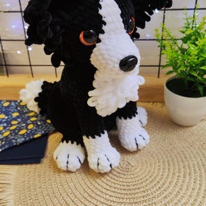 Puede incluir: Un perro de ganchillo hecho a mano en blanco y negro, que se asemeja a un Border Collie. El peluche tiene ojos naranjas y una nariz negra. Est&aacute; sentado sobre un salvamanteles tejido beige, con una peque&ntilde;a planta y una tela estampada en el fondo.