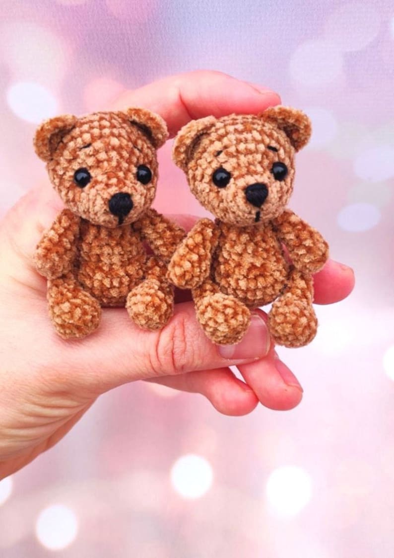 May include: Two small, handmade brown teddy bears with black eyes and noses. The bears are made of a textured material and have a classic teddy bear shape. The bears are held in a hand against a soft pink background.