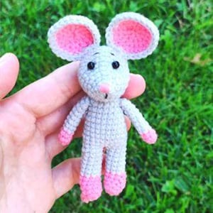 May include: A small, handmade crochet mouse toy featuring a light grey body, large rounded ears with bright pink centres, a small pink nose, and black bead eyes. The mouse has pink accents on its hands and feet, crafted with a visible yarn stitch.