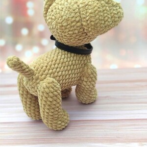 May include: A crocheted plush dog toy with a brown fur texture and a black collar. The dog is standing on a light wood surface.
