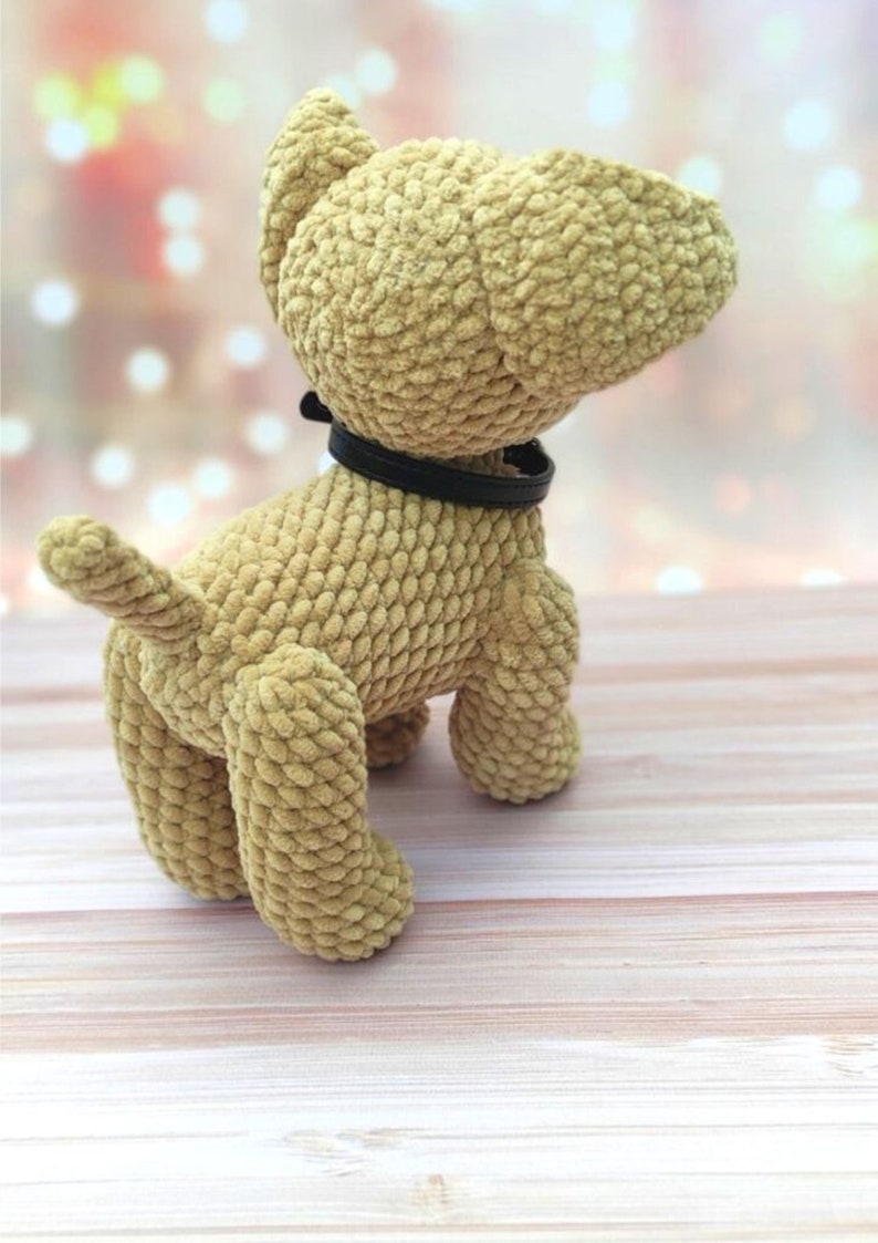 May include: A crocheted plush dog toy with a brown fur texture and a black collar. The dog is standing on a light wood surface.
