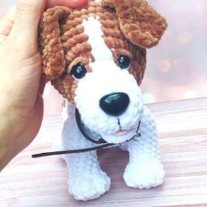 Op de afbeelding: Een gehaakte pluche speelgoedhond met bruin en wit bont. De hond heeft een zwarte neus en bruine ogen. De hond draagt een bruin leren halsband.