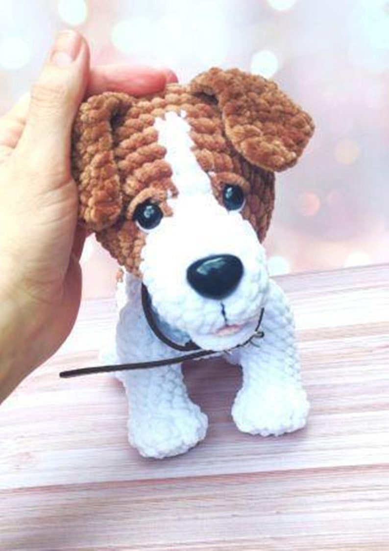 Op de afbeelding: Een gehaakte pluche speelgoedhond met bruin en wit bont. De hond heeft een zwarte neus en bruine ogen. De hond draagt een bruin leren halsband.