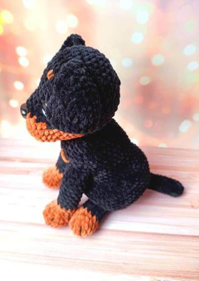 May include: A black and orange crocheted plush toy dog. The dog is sitting on a wooden surface.