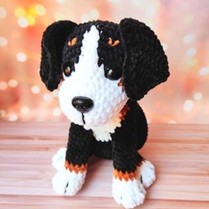 May include: A crocheted Bernese Mountain Dog plush toy with black, white, and brown fur. The toy is sitting on a wooden surface.