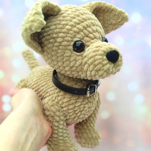 May include: A crocheted plush toy dog with brown fur, black eyes, and a black collar. The dog is sitting on a hand.