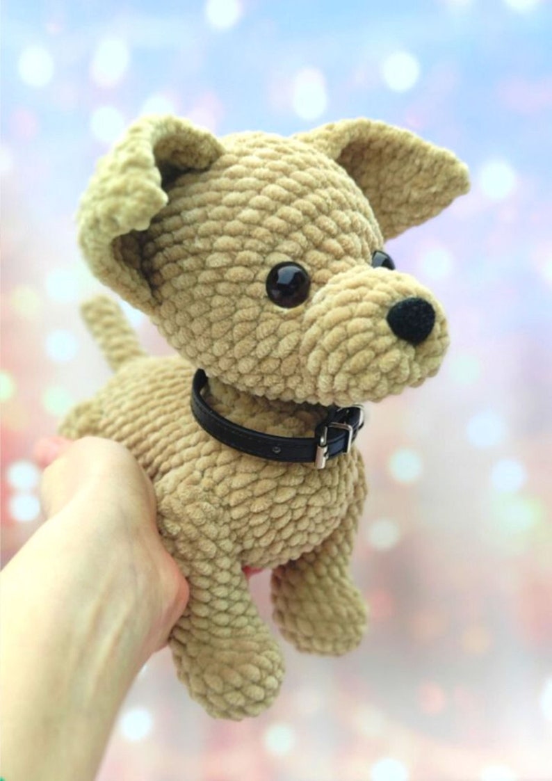 May include: A crocheted plush toy dog with brown fur, black eyes, and a black collar. The dog is sitting on a hand.