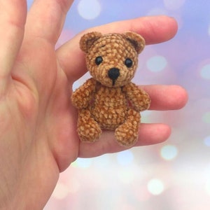 May include: A small, brown crocheted teddy bear with black eyes and a black nose. The bear has a textured appearance and is held in a hand. The background is a soft, blurred mix of colors.