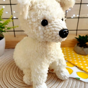 Op de afbeelding: Een handgemaakt, crèmekleurig gehaakt hondenspeeltje met zwarte ogen en neus. Het pluche speelgoed heeft een zachte textuur en staat op een geweven mat. De achtergrond bevat een geel patroon en een kleine plant in een pot.