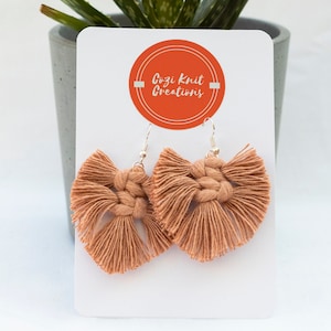 May include: Peach-coloured macrame earrings with silver hooks. The earrings are displayed on a white card with the text "Cozi Knit Creations" in an orange circle. An aloe vera plant is in the background.