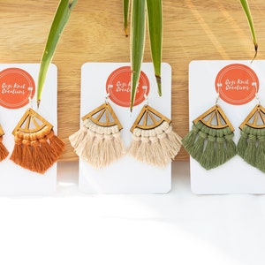 May include: Three pairs of macrame earrings in orange, cream, and green. Each earring has a wooden geometric top and a fringe of knotted cotton. The earrings are displayed on white cards with the text "Goji Knit Creations".