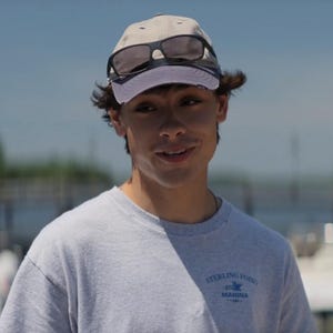 May include: A light gray baseball cap with a curved brim and a pair of black sunglasses resting on the crown. The cap is worn with a light gray crew neck t-shirt featuring the blue text Sterling Point Marina on the upper left chest area.