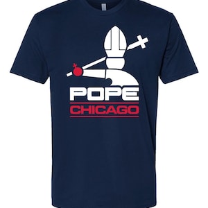 May include: Navy blue t-shirt featuring a white graphic of a pope's hat, cross, and orb, with the word "POPE" above "CHICAGO" in red. The design is centered on the front of the shirt.