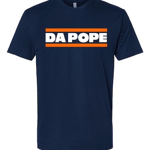 May include: Navy blue t-shirt with the text "DA POPE" in white letters inside an orange rectangle. The t-shirt is a short-sleeved crew neck style.