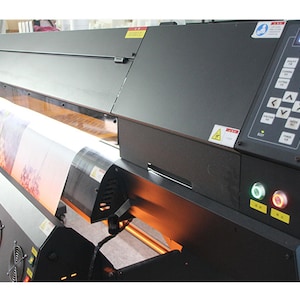 May include: A large format printer with a roll of canvas being fed through the machine. The printer has a control panel with buttons and a display screen that reads "Print Control".