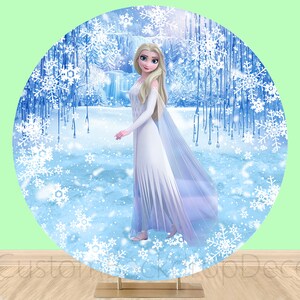 Puede incluir: Un tel&oacute;n de fondo redondo con una escena de invierno con un esquema de color blanco, azul y plateado. Un personaje femenino con cabello largo y rubio y un vestido blanco se encuentra en el centro del tel&oacute;n de fondo. El tel&oacute;n de fondo es perfecto para una fiesta con tem&aacute;tica de Frozen.