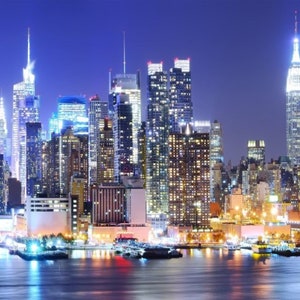 Puede incluir: Una vista nocturna del horizonte de la ciudad de Nueva York con muchos rascacielos iluminados por luces. El Empire State Building es visible en el centro de la imagen.