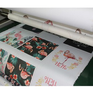 May include: A close-up of a printing press with a roll of white fabric being fed into the machine. The fabric is printed with various designs featuring pink flamingos and tropical plants. Some of the designs include the words "Hello" and "Keep Calm".