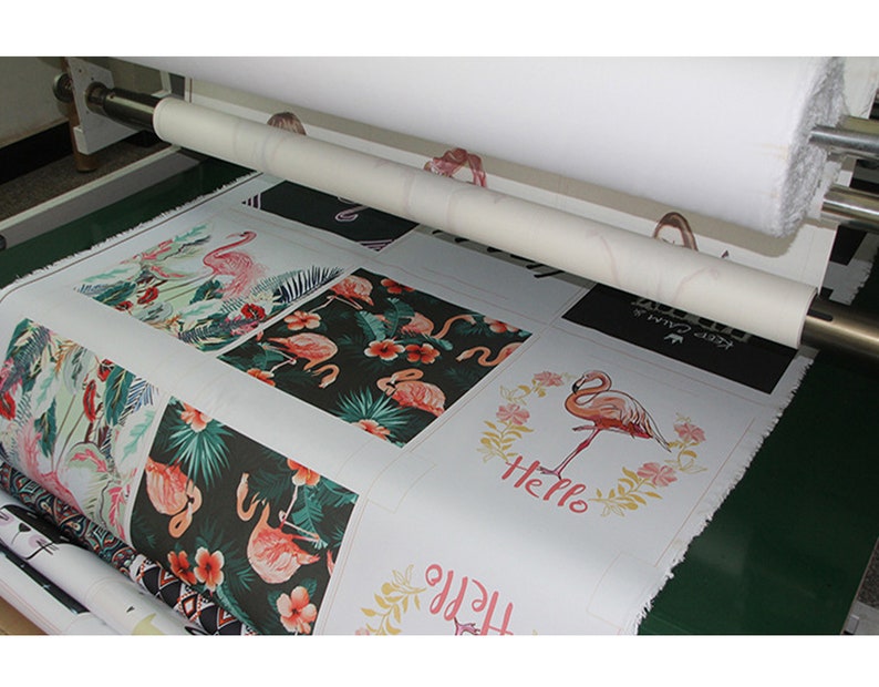 May include: A close-up of a printing press with a roll of white fabric being fed into the machine. The fabric is printed with various designs featuring pink flamingos and tropical plants. Some of the designs include the words "Hello" and "Keep Calm".