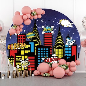 May include: A round backdrop featuring a cityscape with comic book style buildings and speech bubbles with the words "Kaboom!" "Bang!" "Smash!" and "Pow!" The backdrop is blue with white stars and a white cloud. The backdrop is surrounded by pink balloons.