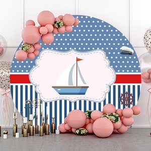 May include: A blue and white striped round backdrop with a red stripe and white polka dots. A white cloud with a sailboat and a red flag is in the center. The backdrop is decorated with pink balloons and greenery.