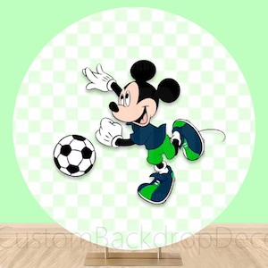 May include: A round backdrop featuring Mickey Mouse in a soccer uniform, reaching for a black and white soccer ball. The background is a light green with a checkered pattern. The backdrop is on a gold stand.
