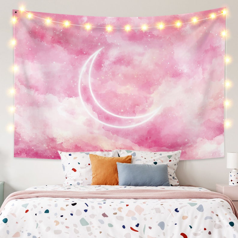 May include: Pink and white tapestry with a crescent moon and stars on a watercolor background. The tapestry is hanging on a wall above a bed with white bedding and colorful pillows.