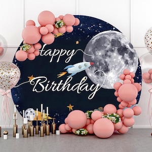 May include: A round, blue and black backdrop with a large white moon, a rocket ship, and the text "happy birthday" in white script. The backdrop is decorated with pink balloons and white flowers.
