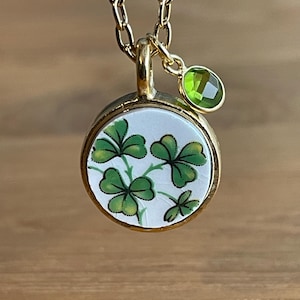 May include: Gold-toned necklace with a round pendant featuring a white background and green shamrock design. A small green gemstone hangs next to the pendant. The chain is a classic link style.