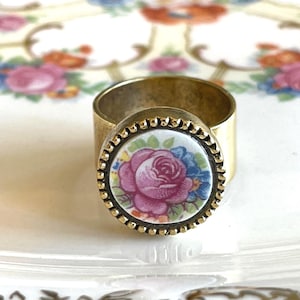 May include: A gold-toned metal ring featuring a circular inset with a white background and a floral design of a large pink rose surrounded by small blue flowers and green leaves. The circular setting has a beaded edge texture around the perimeter.