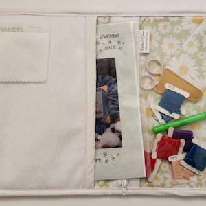 May include: A white fabric project bag with a zipper closure. The bag has a pocket on the front and a clear plastic pocket on the back. The clear pocket contains a cross-stitch pattern, embroidery floss, scissors, and a needle threader. The fabric has a daisy print.