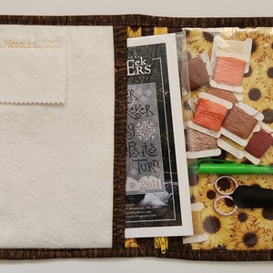 May include: Open craft organizer with a brown floral print exterior and a sunflower print interior. The organizer contains embroidery floss, scissors, a pen, and a cross-stitch pattern. The left side has a white felt pocket with the text "Sera Needles S.O.S."
