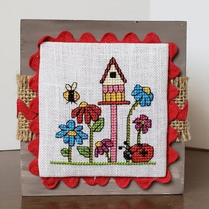 May include: A cross-stitch embroidery on a wooden frame with a red scalloped border. The design features a birdhouse with a pink roof, flowers, a bee, and a ladybug.