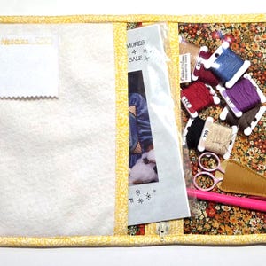 May include: Open sewing kit with a floral print interior and a white, quilted exterior. The kit contains spools of thread in various colors, scissors, a seam ripper, and a pattern. The kit is secured with a zipper and yellow snaps.