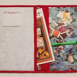 May include: Red fabric project organizer with a clear plastic pocket containing embroidery floss, a needle minder, and a pattern. The organizer has a white felt pocket labeled "Needles" and a white felt pocket with a pattern.