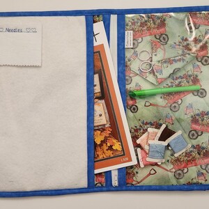 May include: Open sewing organizer with a blue border. The interior features a white felt pocket with "Needles" text, a patterned pocket with red wagons and flags, and a zippered pocket with sewing patterns and spools of thread.