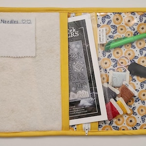 May include: A yellow fabric organizer with a clear plastic pocket. The pocket contains a cross stitch pattern, embroidery floss, scissors, and a needle threader. The organizer has a zipper closure and a white fabric pocket labeled "Needles".