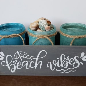 May include: A gray wooden box with the words "beach vibes" painted on the front. The box contains three teal blue glass jars with twine tied around the top. The center jar is filled with seashells.