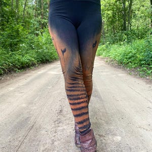 May include: Black leggings with a gradient design transitioning from black to orange, featuring floral and fern patterns. The lower legs showcase a tiger stripe design. The person is wearing brown leather boots with decorative studs.