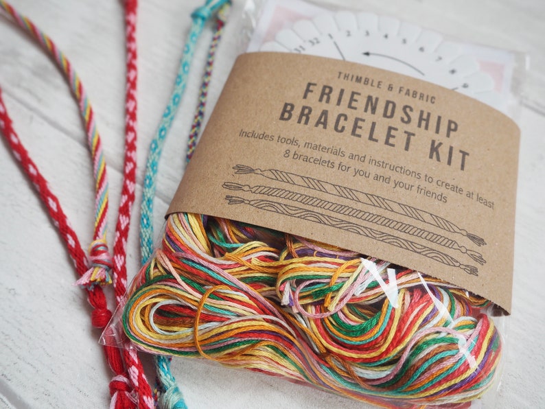 Op de afbeelding: Een knutselset voor het maken van vriendschapsarmbanden. De set bevat kleurrijk borduurgaren, gereedschap en instructies om ten minste 8 armbanden te maken. De verpakking is bruin met de tekst "THIMBLE & FABRIC FRIENDSHIP BRACELET KIT".
